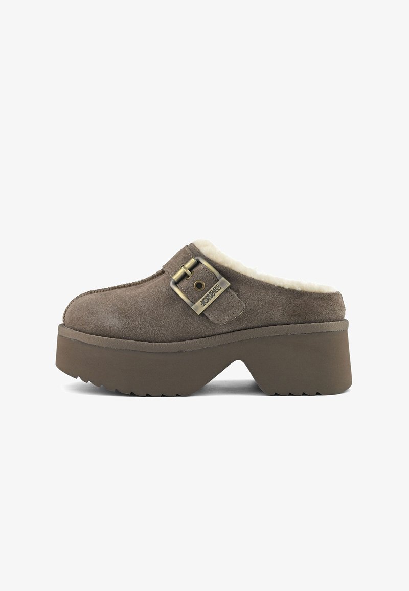 Zueco de ante marrón sin cordones con suela gruesa, forro de piel de oveja y detalle de hebilla metálica ajustable en la parte superior.