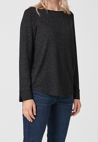 Schwarzes langärmliges Oberteil mit dezenten Glitzerpartikeln, Rundhalsausschnitt und kontrastierenden schwarzen Bündchen. Kombiniert mit blauen Jeans mit einer lockeren Passform.