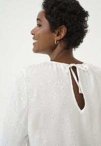 Blouse blanche avec motif texturé, col rond et détail en ouverture à l'arrière noué avec un nœud. Tissu doux et léger, coupe décontractée, manches longues.