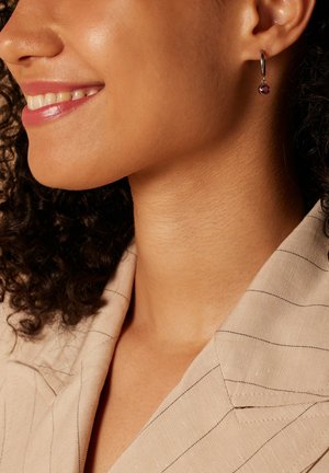 Silberne Creolen mit einem runden lila Edelstein, getragen von einer Person mit lockigem Haar, die ein beigefarbenes Hemd mit Kragen und feinen Streifen trägt.