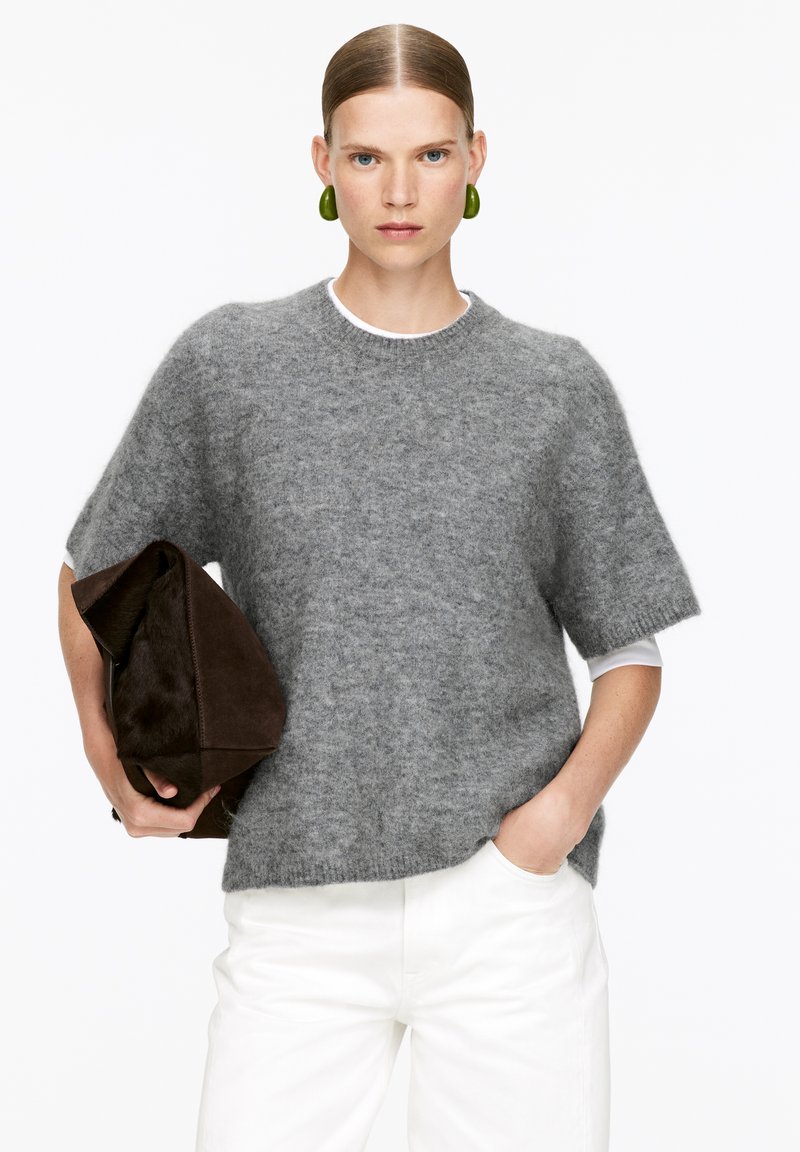 Grauer Strickpullover mit kurzen Ärmeln, Rundhalsausschnitt, kombiniert mit einem weißen Unterhirt. Hält eine dunkelbraune Tasche, trägt grüne Ohrringe.