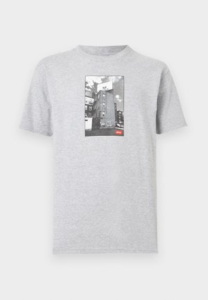 T-shirt gris clair avec une photo en noir et blanc d'un bâtiment et un panneau d'affichage présentant un visage stylisé au-dessus de graffitis, avec une petite étiquette rouge "obey".