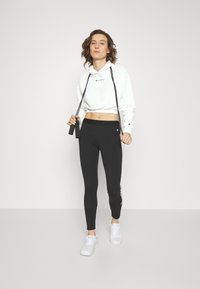 Felpa bianca crop e leggings neri, con dettagli del logo. Il modello tiene una corda per saltare, indossando scarpe da ginnastica bianche. Texture lisce.