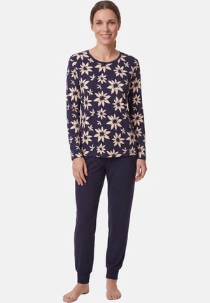 Vrouw staat blootsvoets, draagt een marineblauwe bloemenprint lange mouwen top en bijpassende effen marineblauwe broek tegen een witte achtergrond.