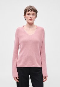 Ροζ πουλόβερ σε σχήμα V, κατασκευασμέ�νο από υλικό σε υφή πλέξη, με μακριά μανίκια και χαλαρή εφαρμογή. Φορεμένο με σκούρα παντελόνια, με μινιμαλιστικό σχέδιο.