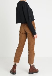 Musta lyhyt neule, vaaleanruskeat corduroy-housut ja ruskeat nauhalliset saappaat. Housuissa näkyvät saumat; saappaissa on keltainen merkki kantapään kohdalla.