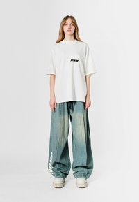 Camisa de gran tamaño blanca con el logo "MWM", combinada con unos vaqueros holgados de un azul claro que presentan un estampado lateral. El modelo lleva calzado de color crema.