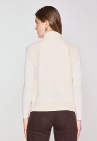 Gilet color crema con colletto alto e design senza maniche. Realizzato in tessuto morbido, presenta una parte posteriore strutturata e orlo a coste.