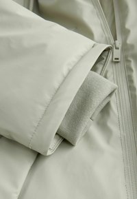 Gaiši zaļa jaka ar gludu, spīdīgu virsmu. Piedurknes galā redzama salocīta pelēka adīta aproce, kas izceļ auduma faktūras kontrastu.