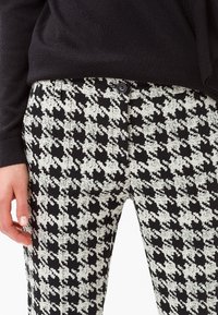Pantalon à motif pied-de-poule noir et blanc avec fermeture à bouton, présentant une texture lisse et une coupe ajustée.