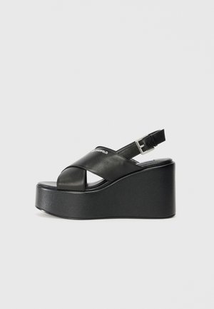 Black platform sandals with a wide, crossed strap design and adjustable ankle buckle. Smooth leather finish and a thick wedge sole.