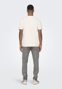 Maglietta a maniche corte di colore chiaro, pantaloni grigi con motivo e sneakers bianche viste da dietro su uno sfondo chiaro.