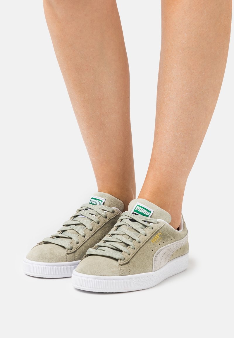 Pieds portant des baskets Puma en suède gris clair avec semelles blanches, lacets noués et logo Puma vert sur la languette, présentés sur fond blanc.