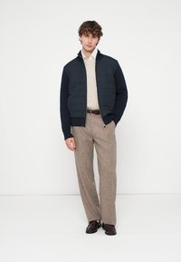 Casaco acolchoado em azul marinho escuro com frente acolchoada, combinado com um suéter creme de malha com tranças e calças de tweed castanho claro, terminado com sapatos escuros.