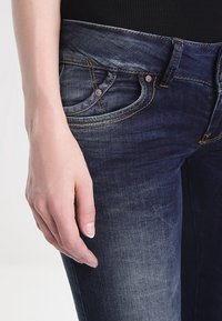 Jeans de mezclilla oscura con un diseño ajustado, un desgaste sutil y costuras en contraste, con bolsillos frontales visibles y detalles en metal plateado.