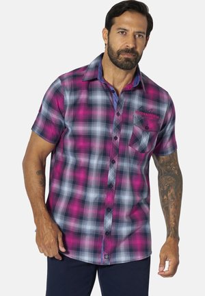 Man wearing a short-sleeve purple and gray plaid button-up shirt with chest pocket and tattooed right arm, standing against white background.