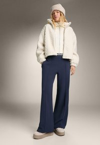 Žena v béžovej pletenej čiapke, krémovej štruktúrovanej bunde na zips, námorníckych nohaviciach s širokými nohavicami a béžových platformových topánkach, stojí s rukami v vreckách.