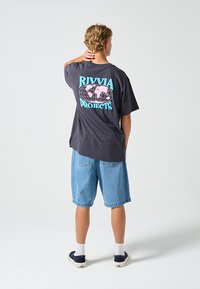 Schwarzes Baumwoll-T-Shirt mit dem Schriftzug "RIVIA PROJECTS" in Blau und Pink sowie einer Globus-Grafik, kombiniert mit blauen Denim-Shorts.