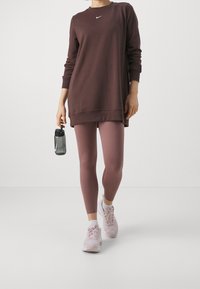 Persona che cammina in leggings viola, felpa oversize di un grigio scuro, scarpe da ginnastica rosa, mentre tiene in mano una bottiglia d'acqua grigia della Nike, su uno sfondo bianco.