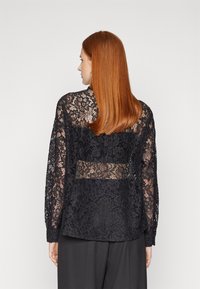 Svart blondebluse med lange ermer, intrikate blomsterdesign og en gjennomsiktig bakpanel. Teksturert blonde med et mykt og delikat utseende.