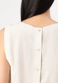 Top senza maniche in lino di un beige chiaro con scollatura rotonda e cinque piccoli bottoni rivestiti in tessuto lungo la schiena. Superficie testurizzata.