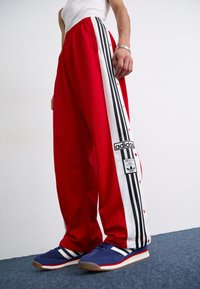 Sarkani sporta bikses ar melniem un baltiem sānu svītrojumiem, ar spiedpogu aizdari un Adidas logo uzlīmi. Materiāls izskatās gluds un viegls.