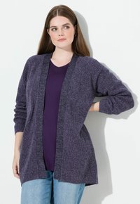 Młoda kobieta z długimi brązowymi włosami nosi fioletowy, dziergany kardigan na ciemnofioletowym topie i jasnoniebieskich jeansach, stojąc na tle białej ściany.