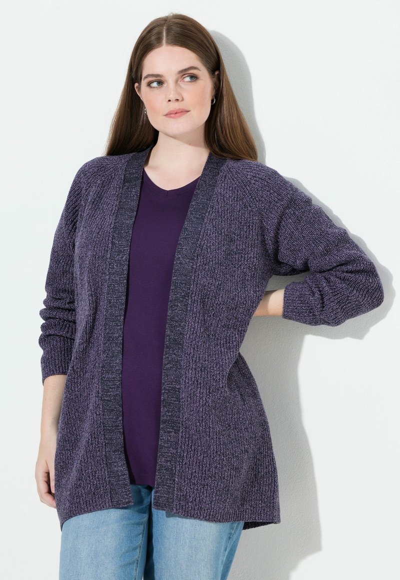 Młoda kobieta z długimi brązowymi włosami nosi fioletowy, dziergany kardigan na ciemnofioletowym topie i jasnoniebieskich jeansach, stojąc na tle białej ściany.