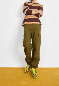 Braun und senfgelb gestreiftes Langarmshirt, olivgrüne Cargo-Hose mit Taschen und leuchtend gelbe Sportschuhe mit schwarzen Akzenten auf gelbem Hintergrund.