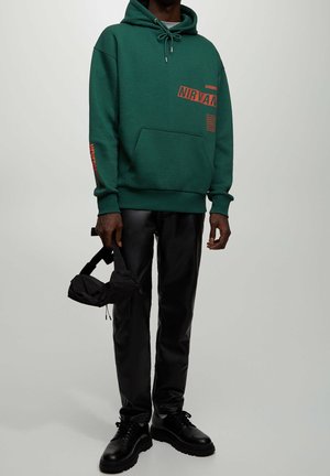 Sweat à capuche vert avec motif "NIRVANA" en orange, poche avant, capuche avec cordon de serrage. Pantalon et chaussures en cuir noir ; petit sac noir tenu à la main.