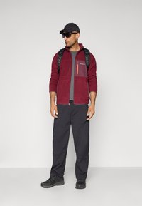 Man wearing a black cap, dark sunglasses, red zip-up jacket, black backpack, gray pants, and black hiking shoes standing against a plain background.