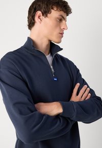 Sudadera con cremallera en azul marino hecha de material suave, con un alto cuello, puños acanalados y un pequeño logo en el pecho.