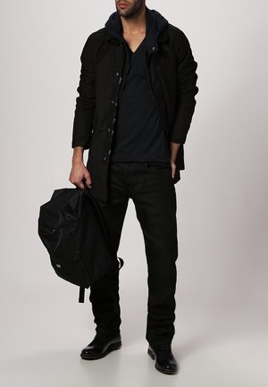 Mand iført sort jakke, sorte bukser, sorte sko, der holder en sort rygsæk ved sin side mod en ensfarvet baggrund.