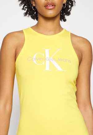 Mujer que lleva una camiseta sin mangas amarilla con el logo "Calvin Klein Jeans" impreso en blanco y lila claro en el pecho.