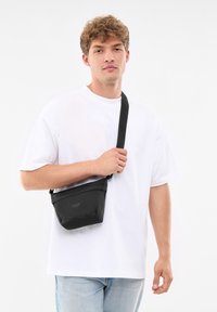 Svart axelväska med en snygg design, dragkedjestängning och justerbar rem, bärs över kroppen över en vit t-shirt och jeans.