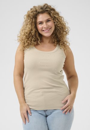 Beige, geripptes Tanktop mit U-Ausschnitt, das auf der Vorderseite kleine klare Verzierungen in einem Muster zeigt. Dazu werden hellblaue Jeans getragen.