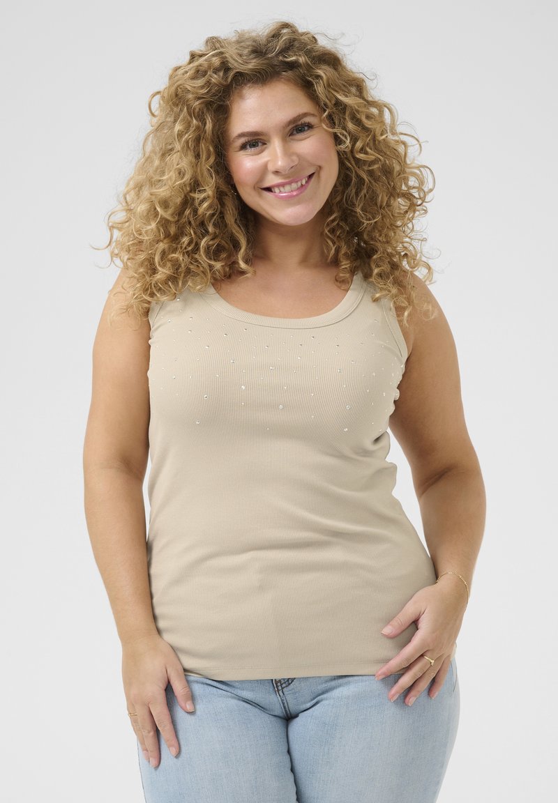 Débardeur beige à côtes avec un décolleté en scoop, présentant de petites ornements transparents disposés en motif sur l'avant. Porté avec un jean bleu clair.