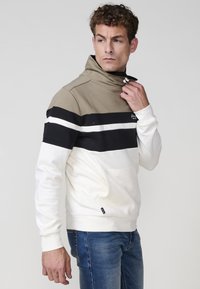 Mikina s farebným blokovaním v béžovej, čiernej a bielej farbe. Vysoký golier, dlhé rukávy a predná kapsa. Mäkka textúra, voľný strih.