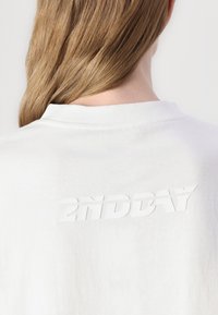 Biela bavlnená tričko s vyvýšeným, textúrovaným potlačou "2NDDAY" na chrbte. Jednoduchý okrúhly výstrih a mäkká textúra.