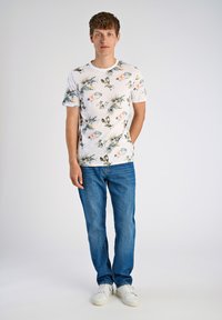 Miestna krátkymi rukávmi biela tričko s farebným kvetinovým a listovým vzorom, spárované s modrými džínsy a bielymi teniskami.