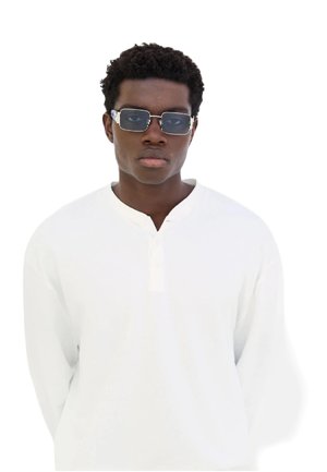 Occhiali da sole con forma rettangolare e lenti scure. Montatura trasparente con accenti metallici. Indossati da una persona con una camicia bianca a maniche lunghe.