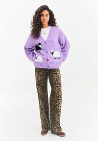 Kobieta w lawendowym kardiganie z wzorem owiec, w spodniach w panterkę, brązowych butach, stojąca na tle jednolitej białej ściany.