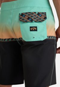 Shorts de bain avec un dégradé allant de teal à noir, dotés d'une poche arrière à motifs et de coutures contrastées. Comprend un patch logo.
