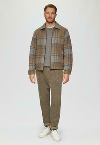Bruine en grijze geblokte jas met een kraag, gedragen over een grijze trui en olijfgroene corduroy broek, aangevuld met witte sneakers.