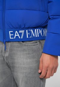 Kék puffertakaró dzseki bordázott kék derékpánttal, amelyen fehér "EA7 EMPORIO" felirat található, világosszürke farmernadrággal párosítva.