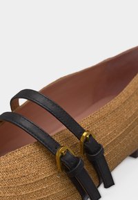 Πλεγμένο ψάθινο επίπεδο παπούτσι με δύο μαύρα δερμάτινα λουριά στερεωμένα με χρυσές αγκράφες στην άνω άκρη.