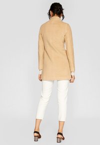 Beige wool blend coat with a fitted silhouette, stand collar, and back seam detailing, paired with white cropped trousers and high-heeled sandals.