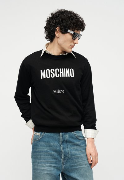 Homme aux cheveux bouclés portant un pull noir Moschino Milano, un jean bleu, une chemise à col blanc et des lunettes de soleil noires Moschino, regardant vers la droite.