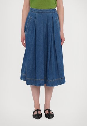 Femme portant une jupe midi en jean plissée avec des poches latérales, accompagnée de chaussures plates tressées noires à bouts pointus et d'un haut vert.