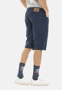 Shorts bleu marine avec une coupe droite, dotés de poches arrière et d'une patch en cuir décoratif sur la taille. Portés avec des baskets blanches et des chaussettes grises.